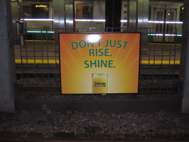 A yellow and orange brightly colored sign the features a glass of orange juice. Makes one a bit thirsty. Seems to be in a metro station.