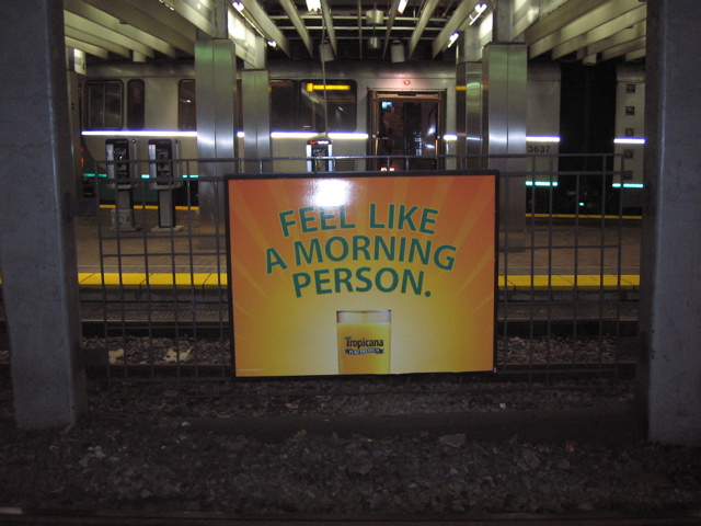 A yellow and orange brightly colored sign the features a glass of orange juice. Makes one a bit thirsty. Seems to be in a metro station.