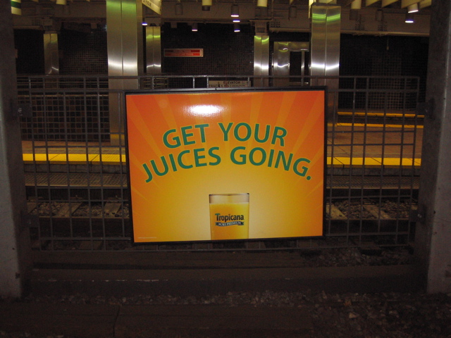 A yellow and orange brightly colored sign the features a glass of orange juice. Makes one a bit thirsty. Seems to be in a metro station.
